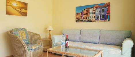 A living room with a wicker chair, a sofa, a glass coffee table, and two paintings on the wall.