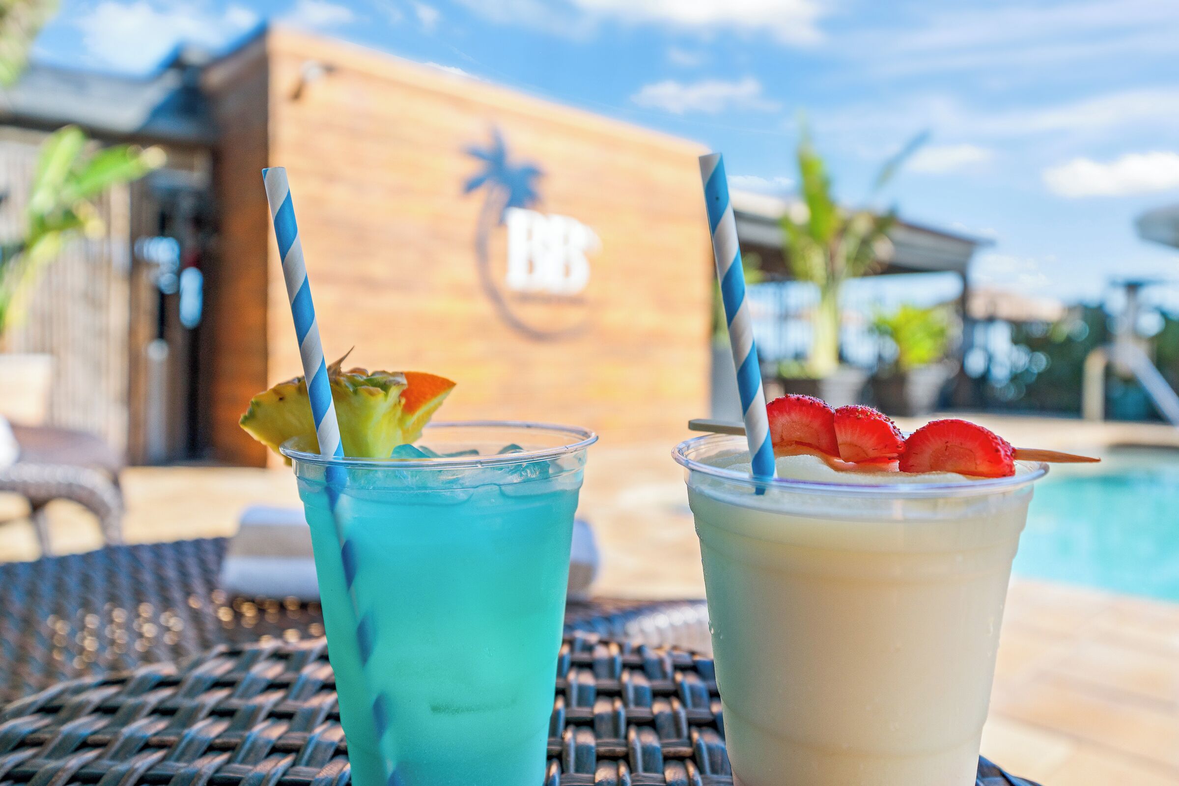 Two drinks with straws and fruit garnishes on a table by a poolside.