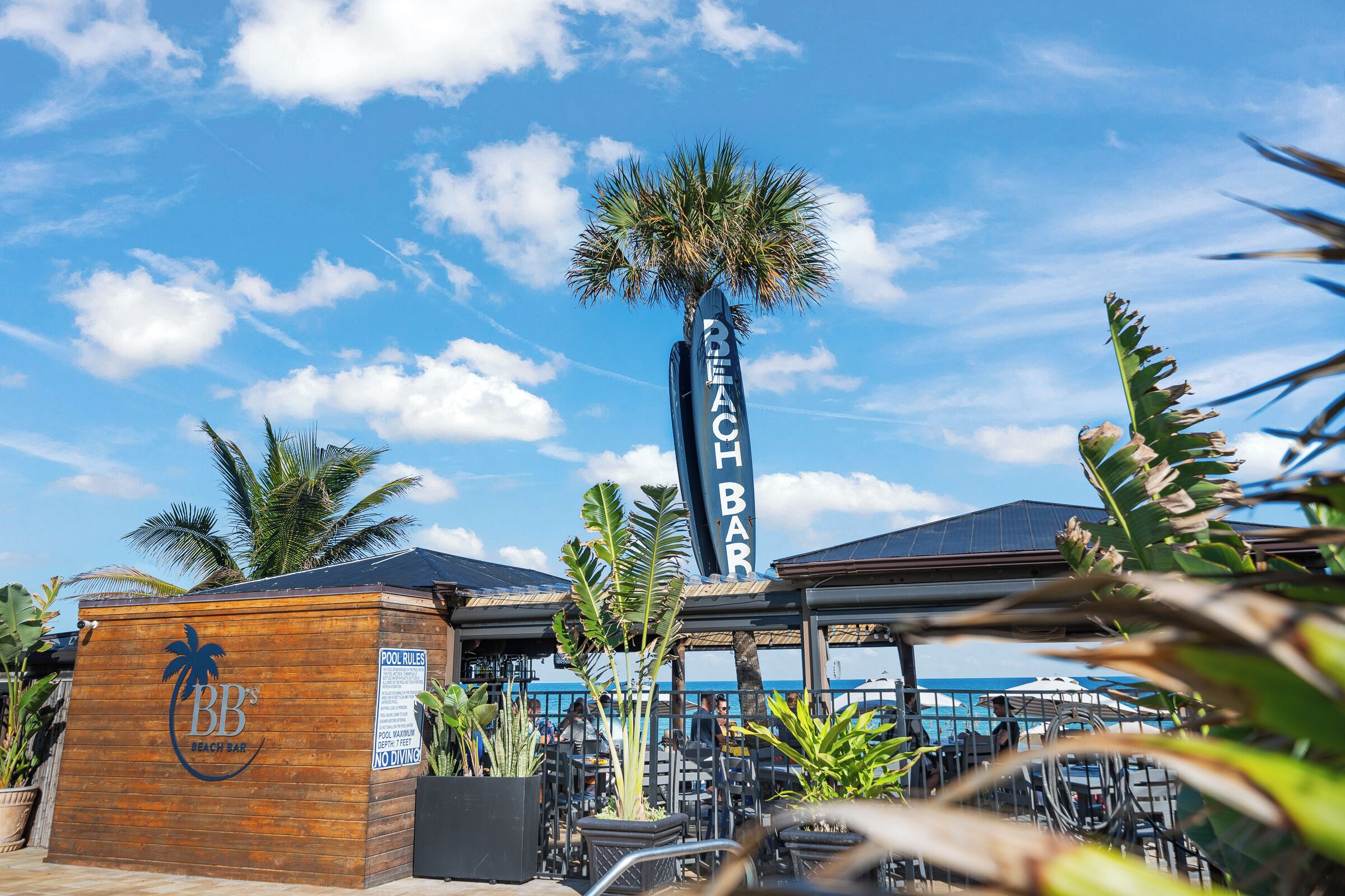 Een strandbar met een houten bord, terras en palmbomen.