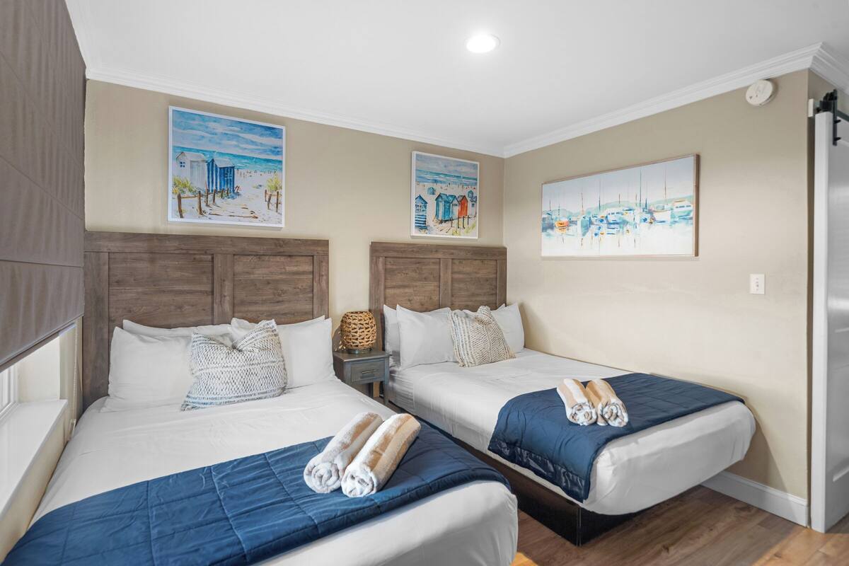 Habitación de hotel con dos camas, paredes color beige y obras de arte que muestran escenas de la costa.