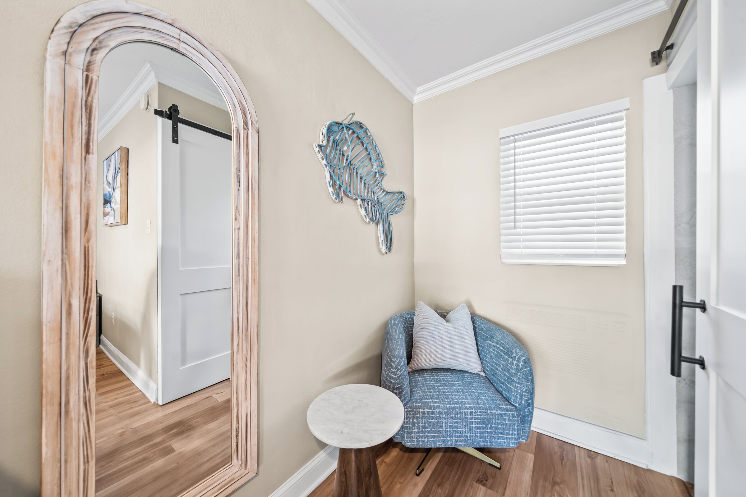 A hallway with a wooden floor, a white door, a blue patterned armchair, a small white table, and a wall-mounted sculpture.