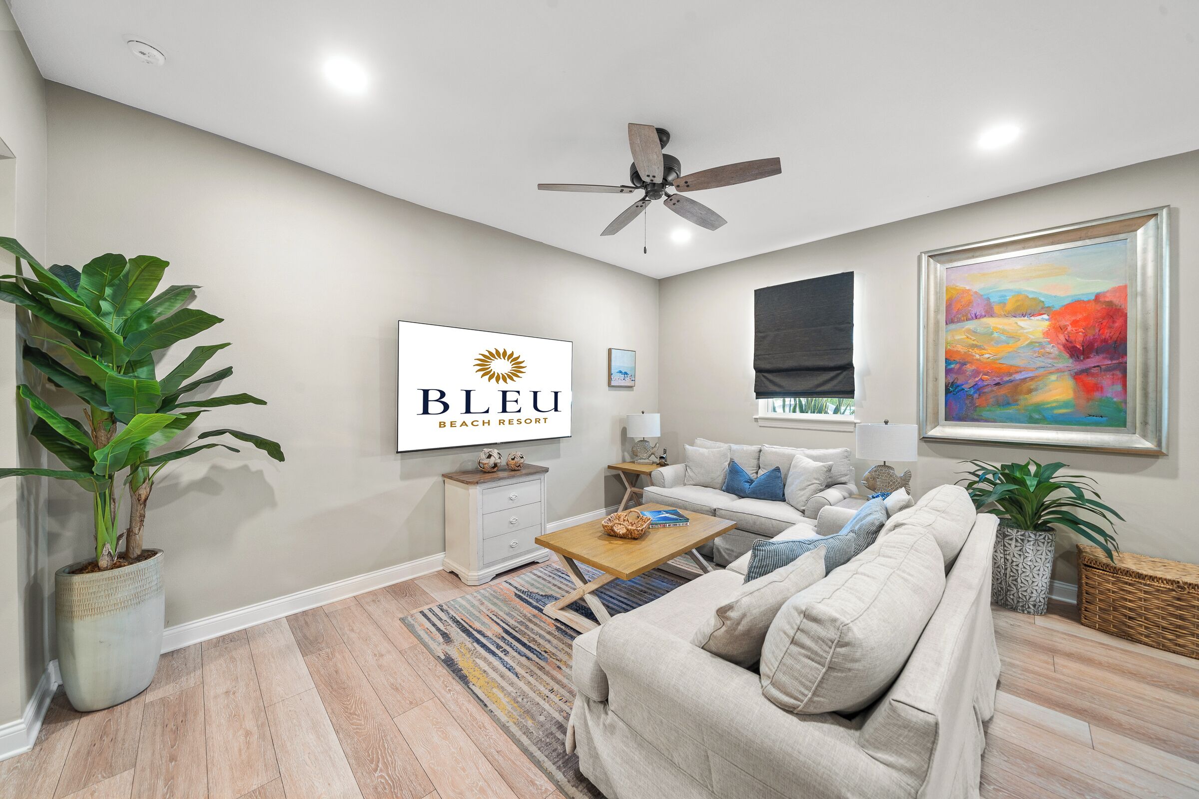 Un salon avec un canapé beige, une table basse, un ventilateur de plafond, un grand tableau et une enseigne sur laquelle on peut lire : « BLEU BEACH RESORT ».