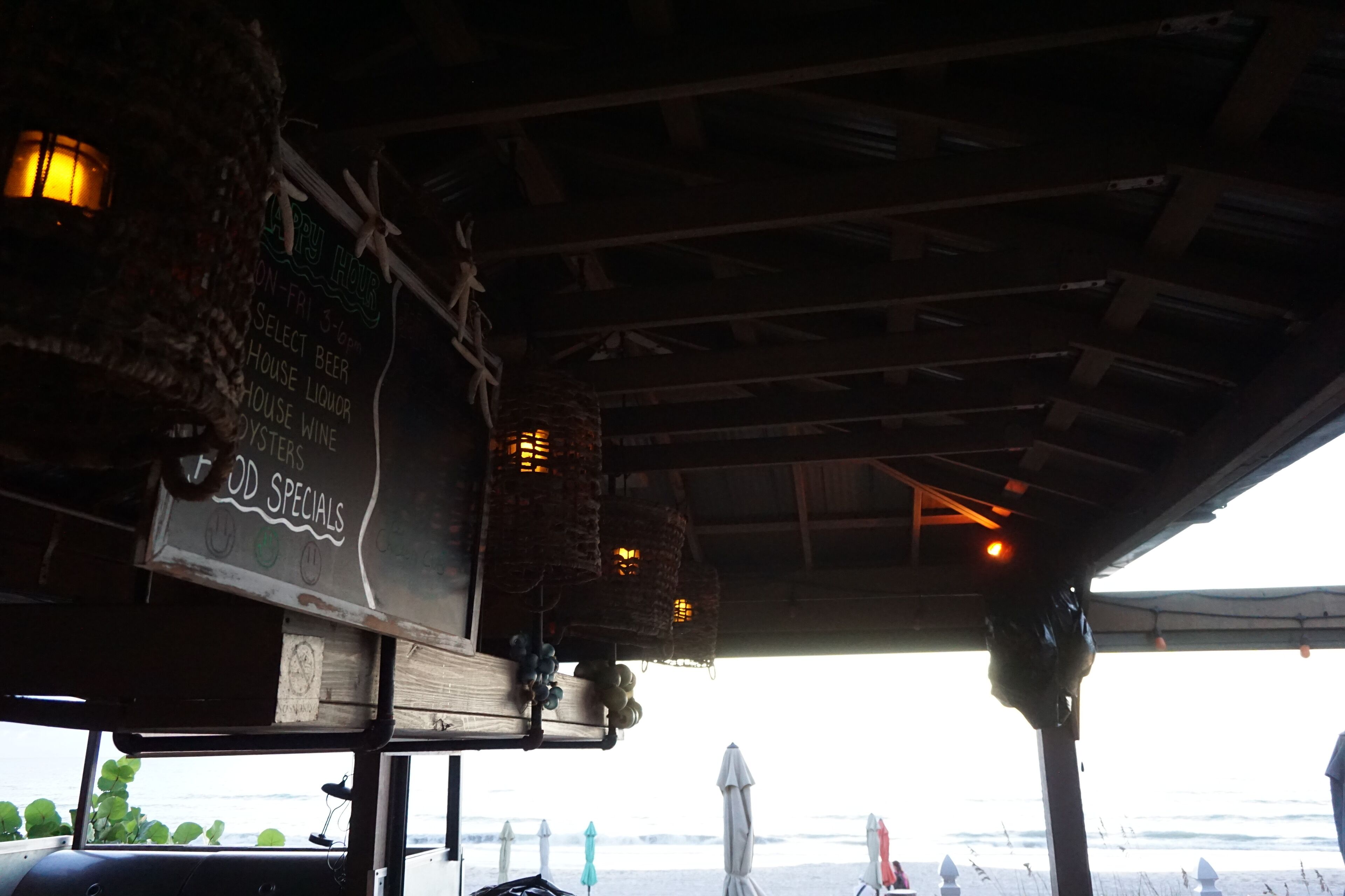 Un ristorante sulla spiaggia con soffitto in legno, cesti fioriti appesi e una lavagna con i piatti del giorno.