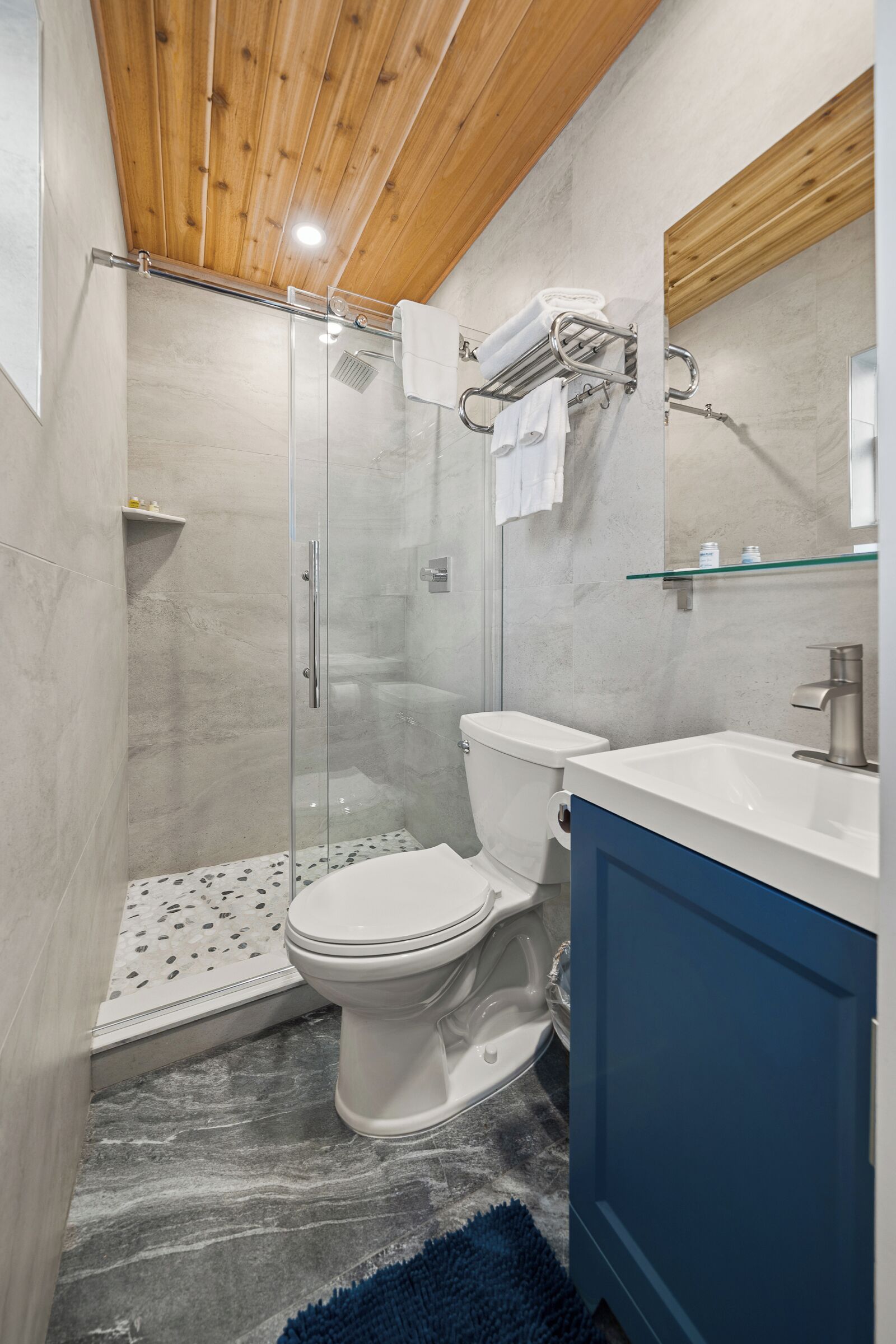 Un baño con cabina de ducha de vidrio, inodoro blanco, mueble azul y repisa de vidrio sobre el lavamanos.