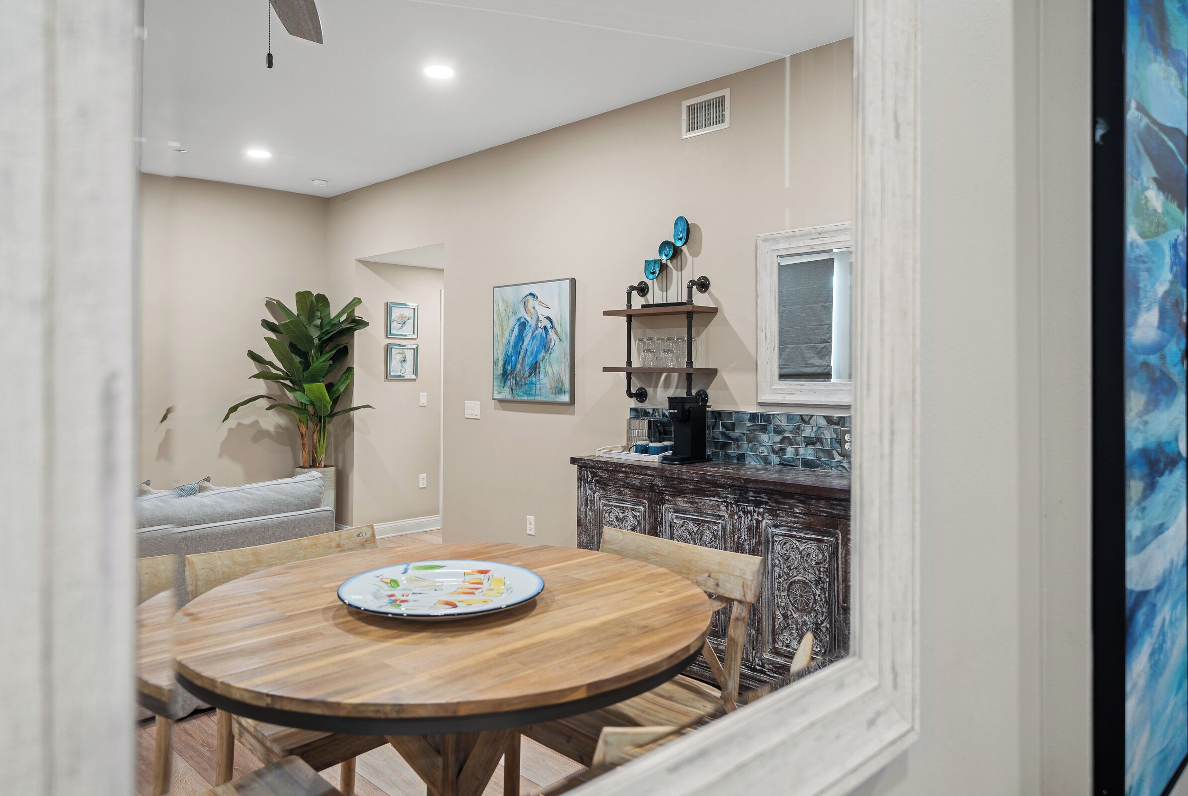 A dining area with a round wooden table, chairs, a potted plant, and a kitchen area with a tiled backsplash and a coffee machine.