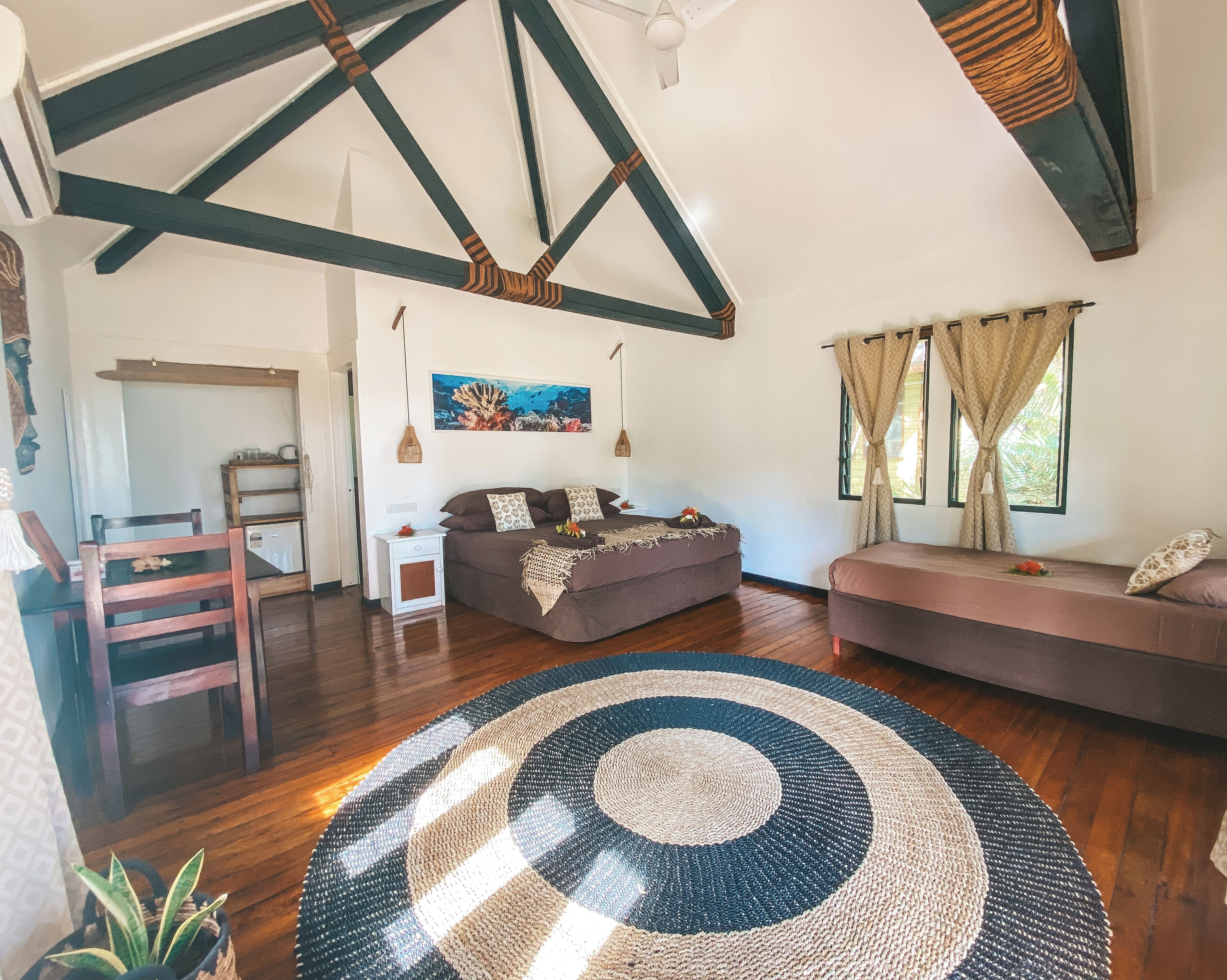 A spacious room with a high ceiling, wooden beams, a large bed, a sofa, a dining area, and a view of the outdoors.
