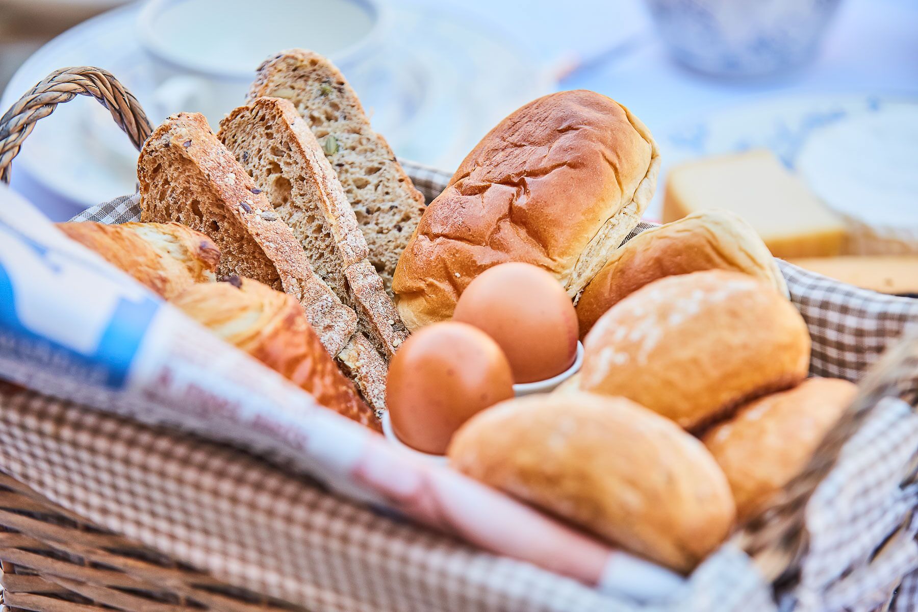 A basket containing various types of bread, including whole grain and baguette, along with a few eggs.