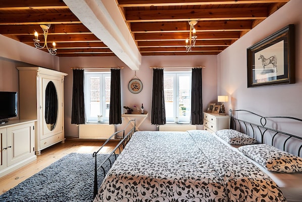 Un dormitorio con techo de madera, una cama con ropa de cama estampada, un aparador, un televisor y dos ventanas con cortinas.
