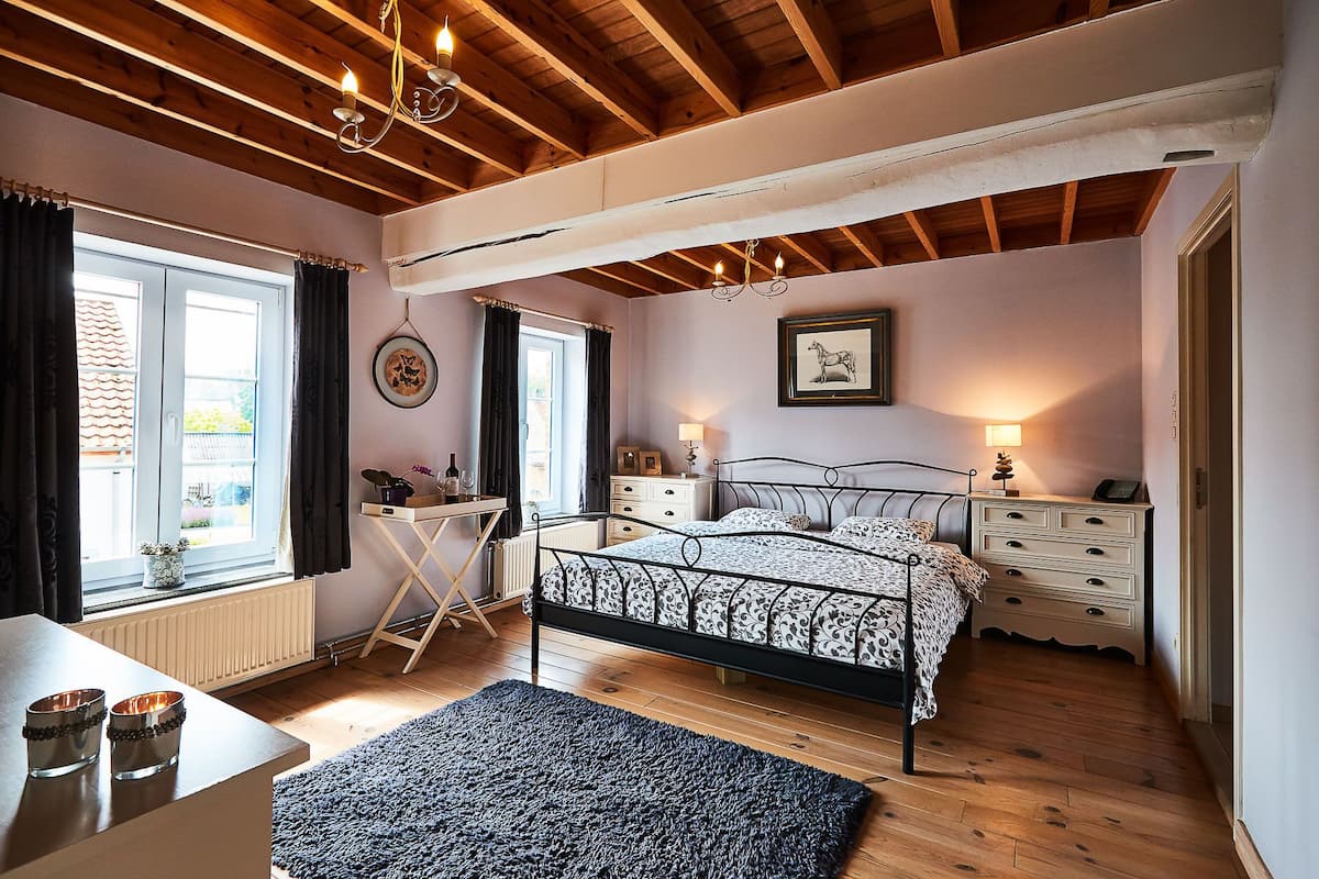 Quarto com cama, mesas de cabeceira, radiador, janela com cortinas, lustre e quadro na parede.