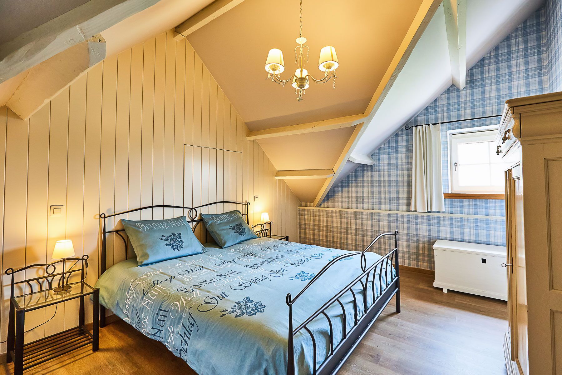 A bedroom with a bed, bedside tables, a chandelier, a window with curtains, and a white dresser.
