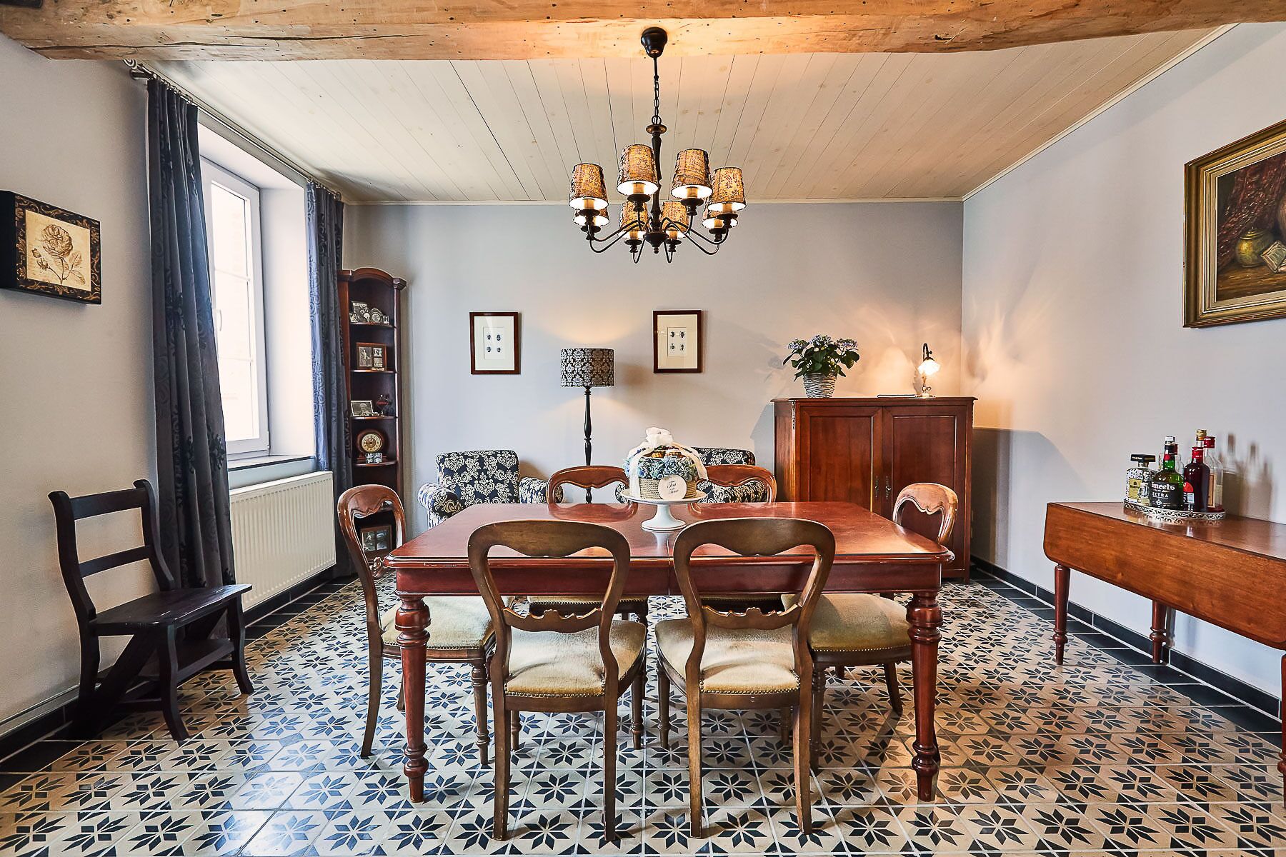 A dining room with a wooden table, chairs, a chandelier, a cabinet, and a patterned floor.
