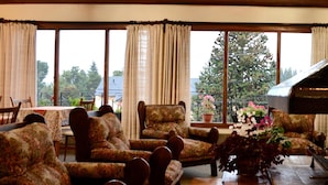A room with floral-patterned armchairs, a wooden table, and large windows with curtains. - Hosteria Güemes (San Carlos de Bariloche)