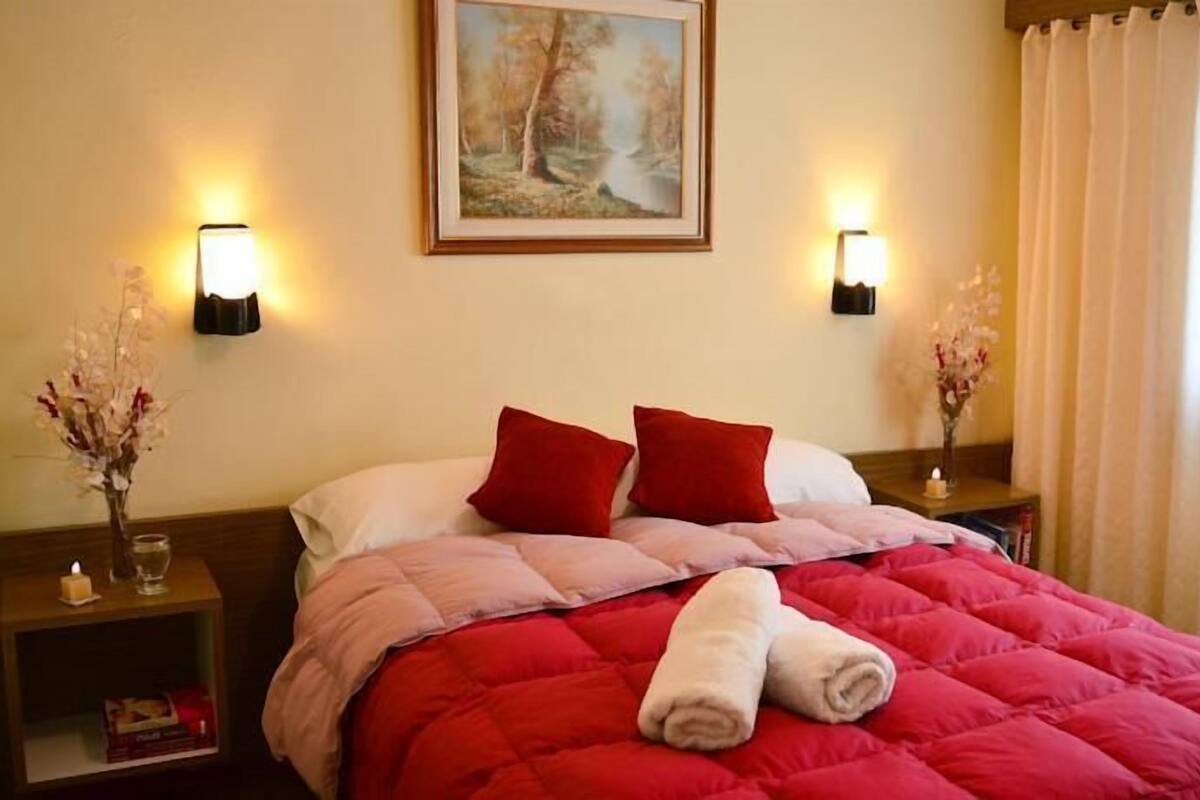 Un lit bien fait, avec une literie rouge et blanche, deux oreillers rouges et des serviettes enroulées posées sur le lit. Il y a deux tables de chevet, chacune avec une lampe et un vase de fleurs. Un tableau est accroché au mur, au-dessus du lit.
