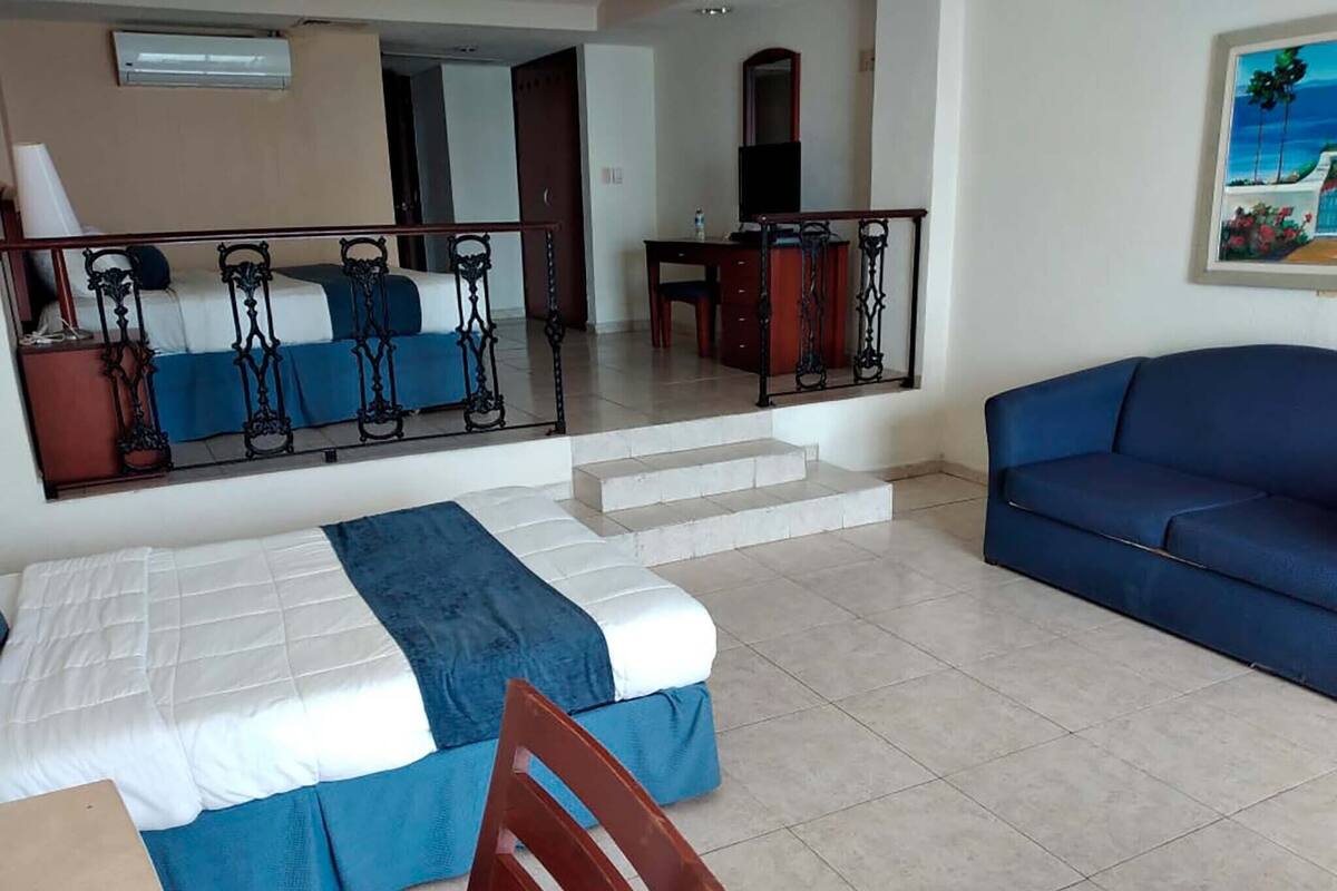 Une chambre d’hôtel avec un lit, un canapé bleu, une chaise en bois, un bureau et un escalier menant à un balcon.
