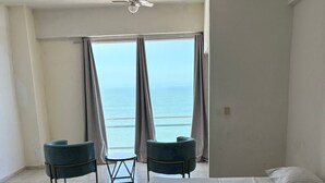 Habitación de hotel con una cama, dos sillas, una mesita, un ventilador de techo y vista al océano.