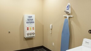 A laundry room with a Whirlpool washer, a blue ironing board, a wall-mounted iron, and a sign listing soap, bleach, and softeners. - Comfort Inn & Suites West Artesia (Artesia)