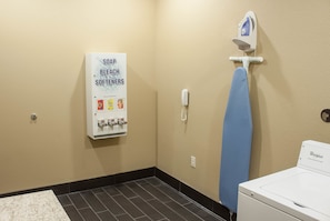 A laundry room with a Whirlpool washer, a blue ironing board, a wall-mounted iron, and a sign listing soap, bleach, and softeners. - Comfort Inn & Suites West Artesia (Artesia)