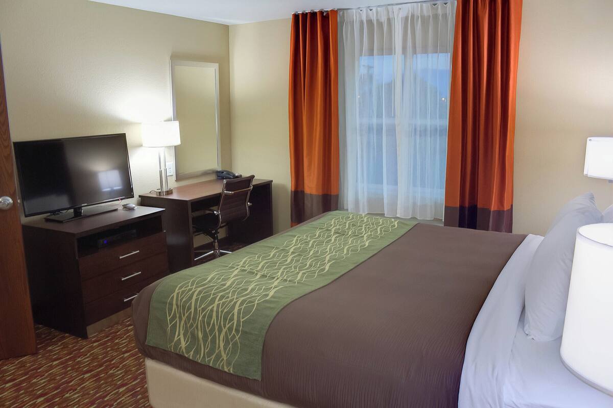 Habitación de hotel con cama, escritorio con computadora, televisor y ventana con cortinas.