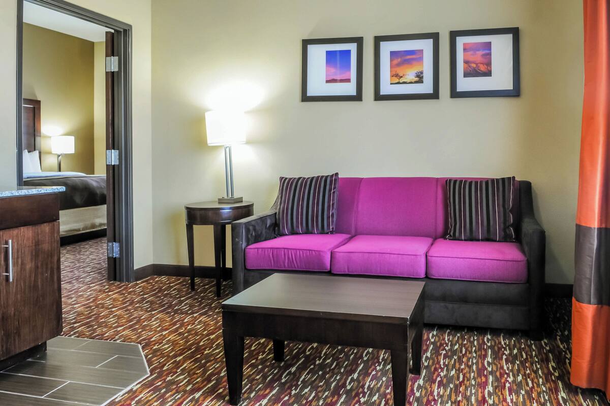 Habitación de hotel con sofá morado, una mesa de centro de madera, una lámpara y una cama con mesita de noche. En la pared, hay tres cuadros enmarcados.