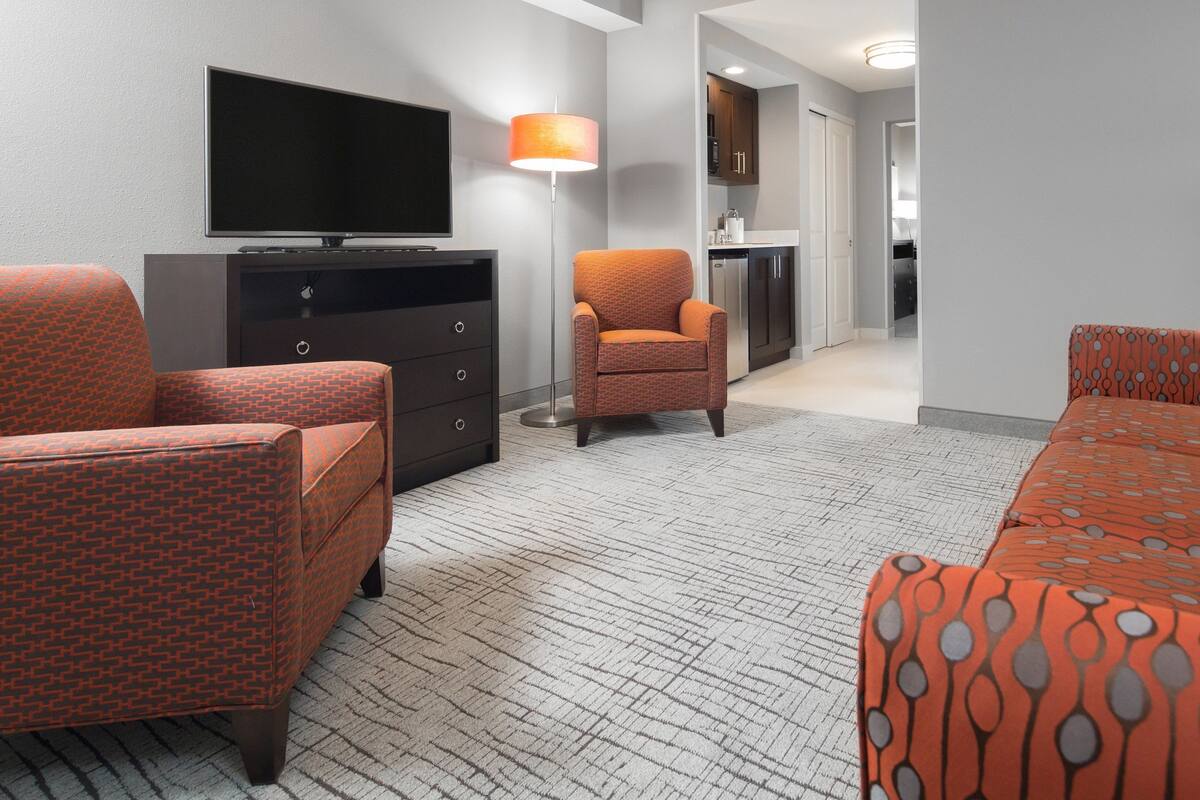 Ein Hotelzimmer mit Fernseher, Sofa, Sessel, Lampe und einer Küchenzeile.