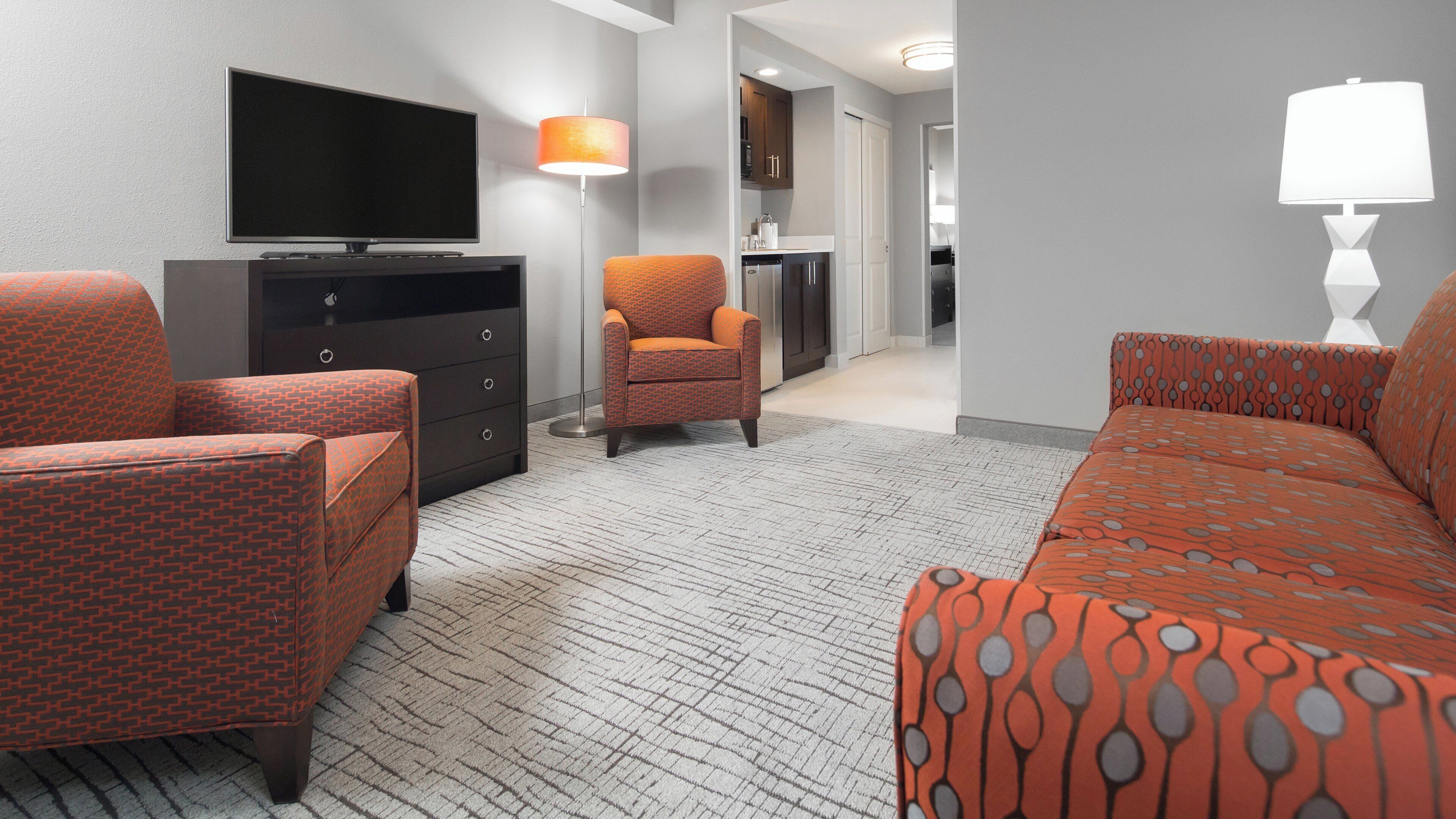 A hotel room with a television, a sofa, an armchair, a lamp, and a kitchenette.
