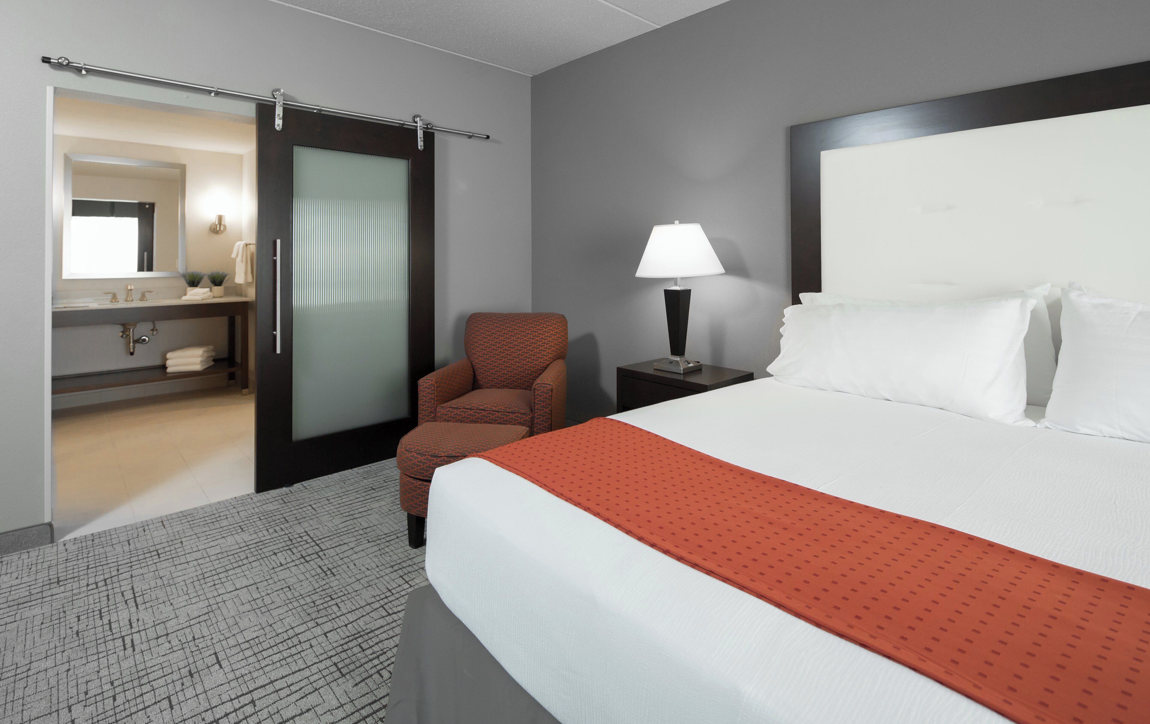 Quarto de hotel com cama, cadeira, mesa de cabeceira, luminária, banheiro com espelho e porta de correr.