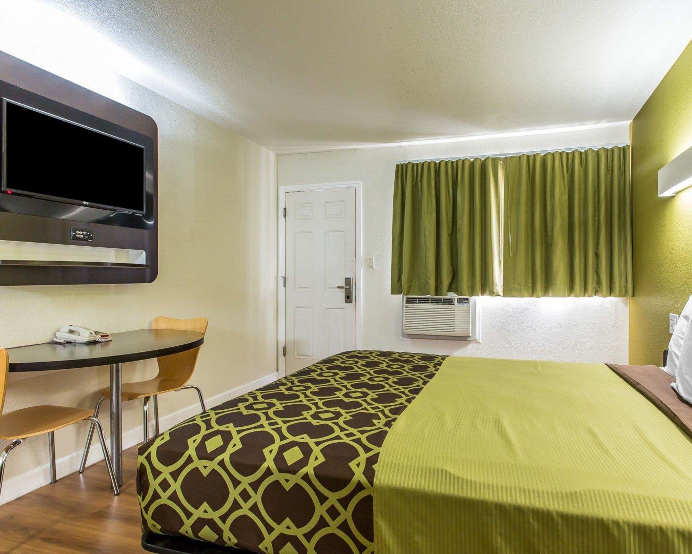 Una camera d'albergo con un letto, una scrivania, una sedia, una televisione, una porta e una finestra con tende.