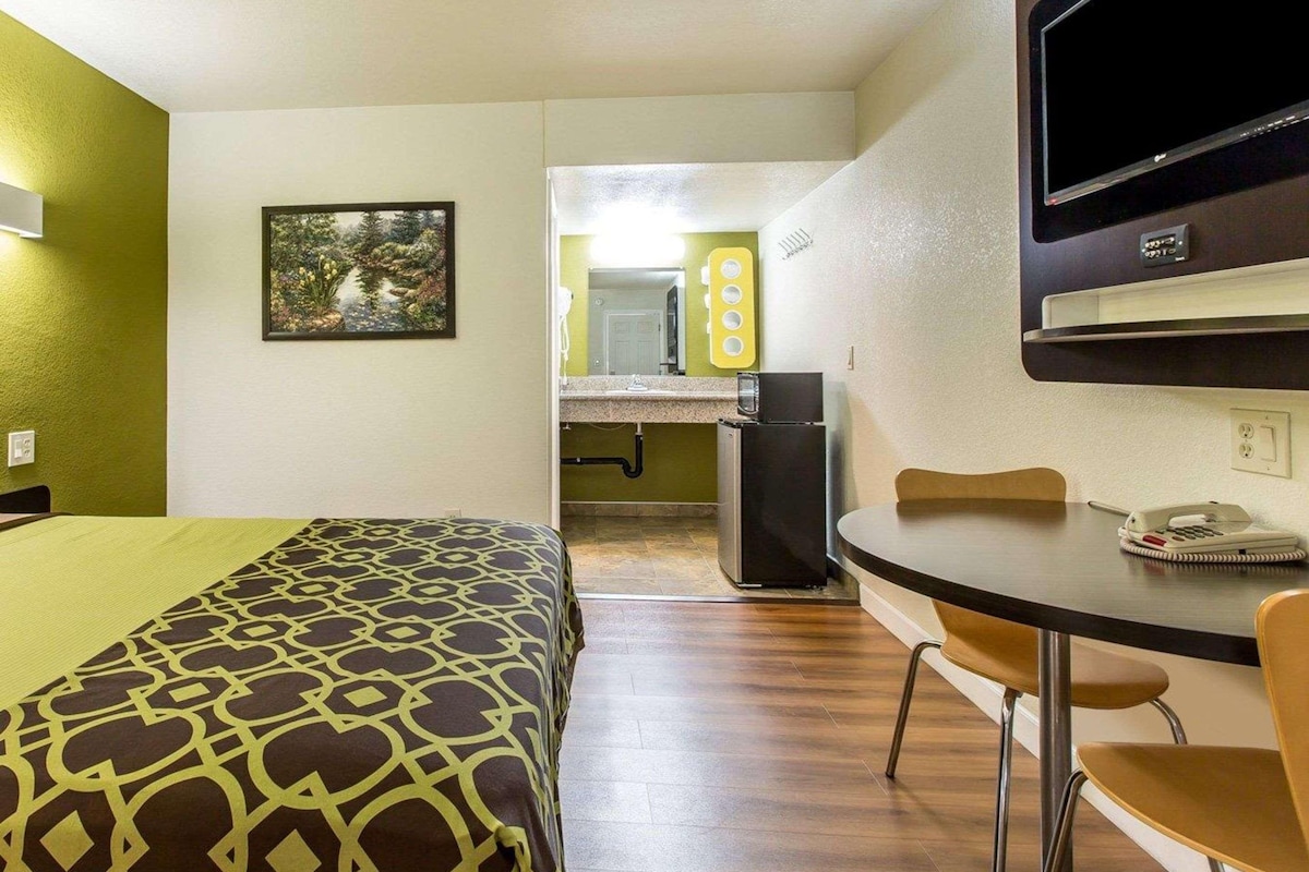 A hotel room with a bed, a desk, a chair, a TV, and a bathroom visible in the background.