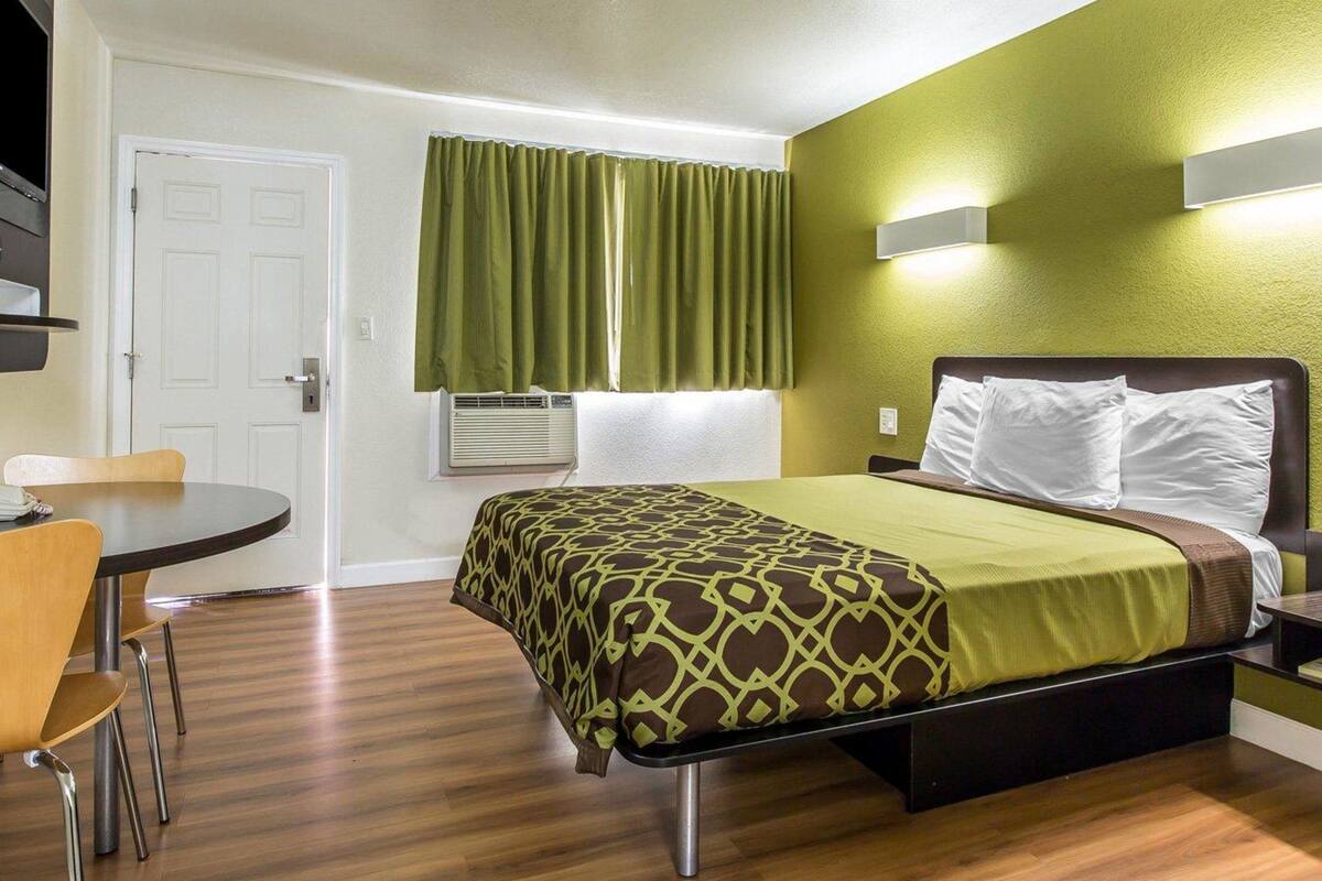 Quarto de hotel pequeno com cama, mesa de cabeceira e uma pequena mesa redonda com cadeiras.