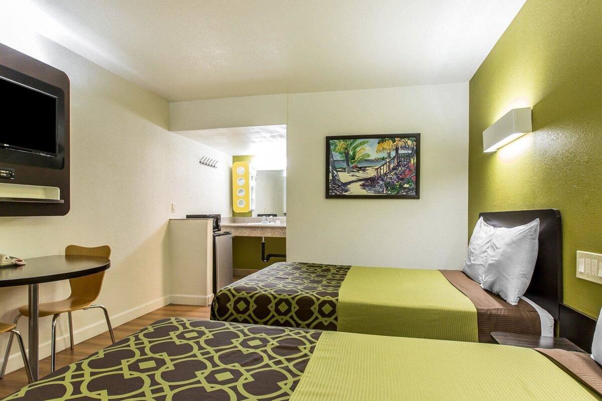 Quarto de hotel com duas camas, uma escrivaninha, uma cadeira, uma televisão e um quadro na parede.