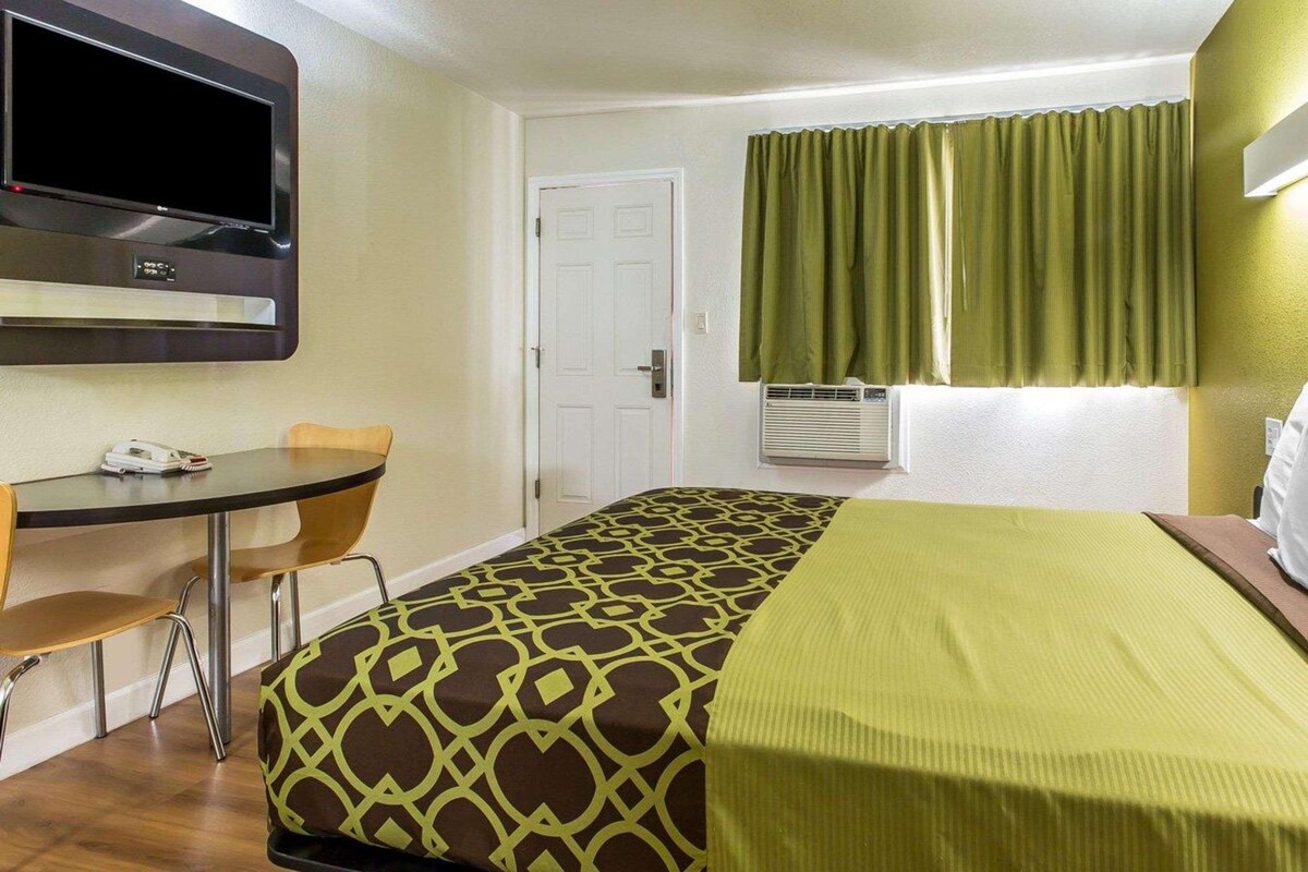 A hotel room with a bed, a desk with a chair, a television, a door, and a window with curtains.