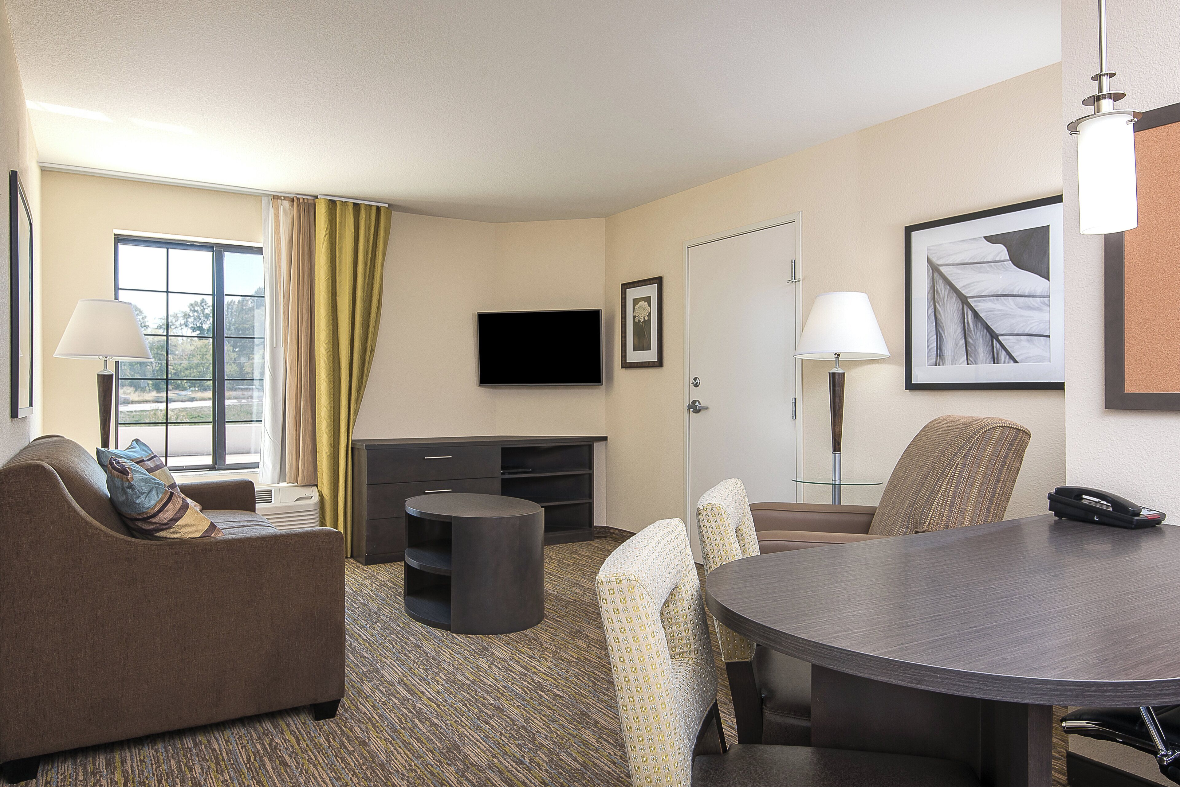 A hotel room with a sofa, a small table, a larger dining table, a flat-screen TV, a lamp, and a framed picture on the wall.