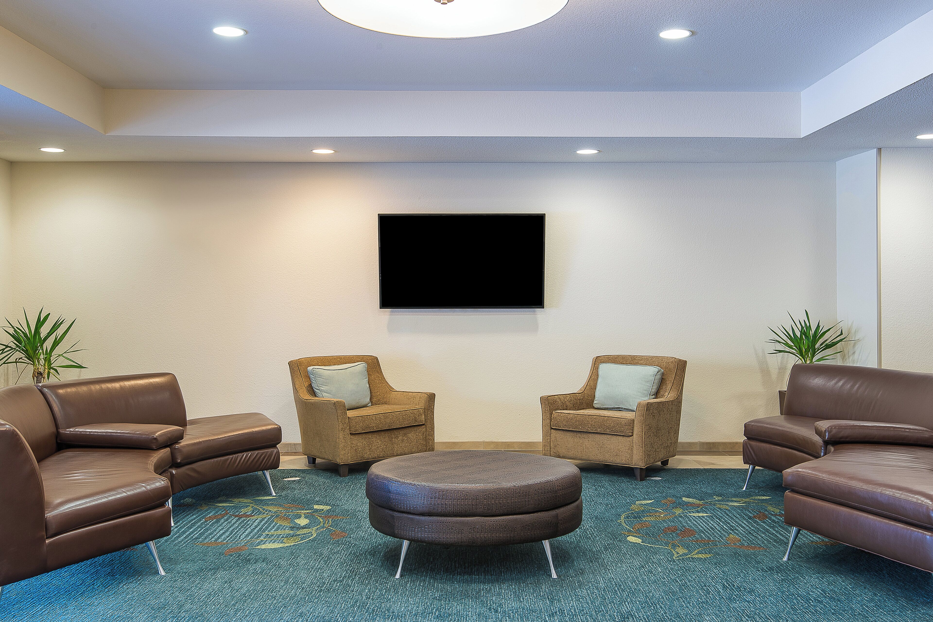 A lobby with a flat-screen TV mounted on the wall, two armchairs with cushions, a round ottoman, and two potted plants.