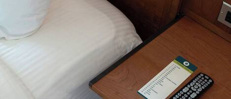 A hotel room with a bed, a wooden nightstand, a remote control, and a card with text.