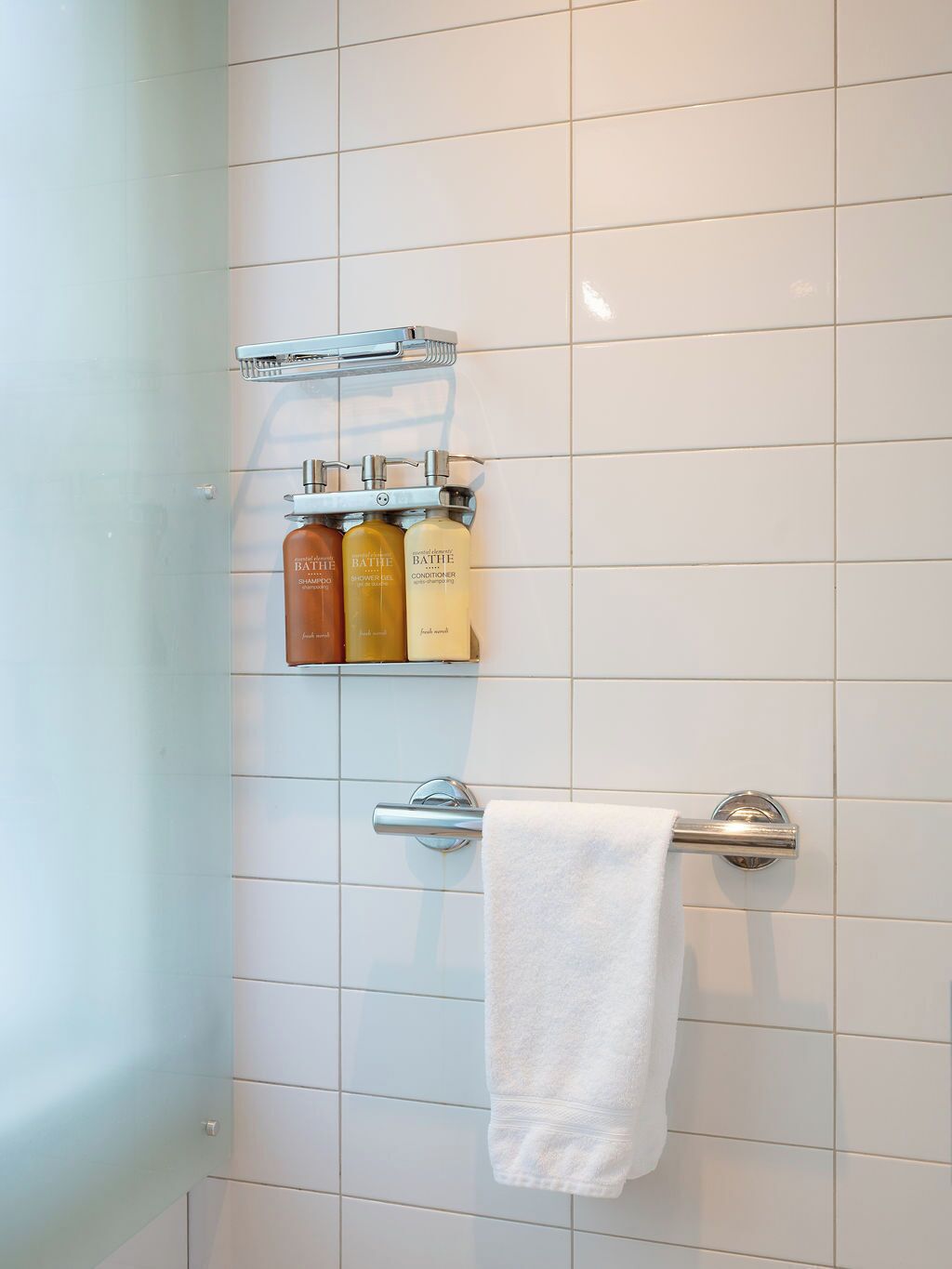 Ein Badezimmer mit weißen Fliesen, einem Handtuchhalter mit einem weißen Handtuch und drei Badezimmerartikeln auf einem Regal.