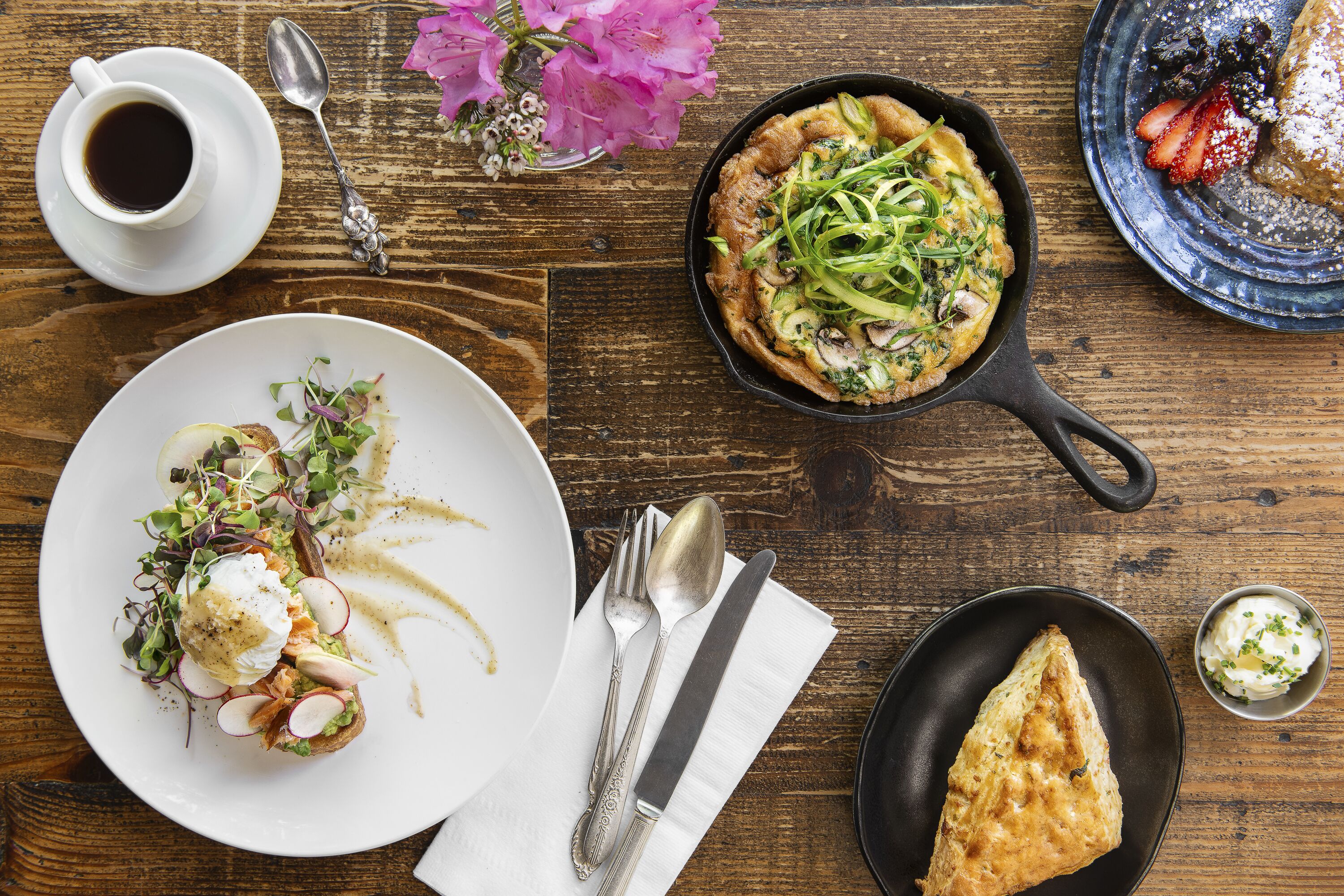 A wooden table with various dishes, including a plate with a poached egg and greens, a skillet with a baked dish, and a cup of coffee.