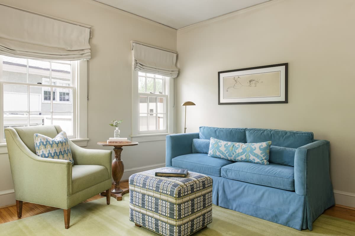 A living room with a light green armchair, a blue sofa, and a patterned ottoman. There are two windows with white curtains, a framed picture on the wall, and a small table with a vase.