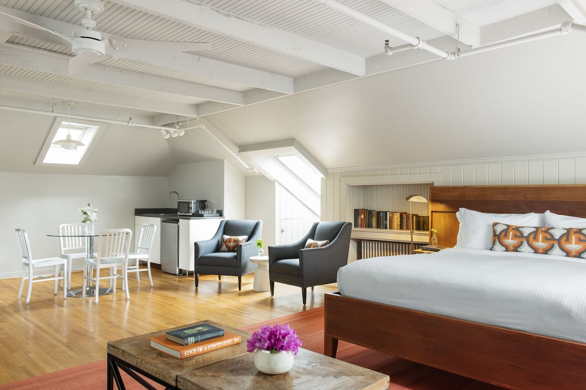 A modern bedroom with a large bed, a desk, and two chairs. There is a skylight above the desk and a bookshelf against the wall.