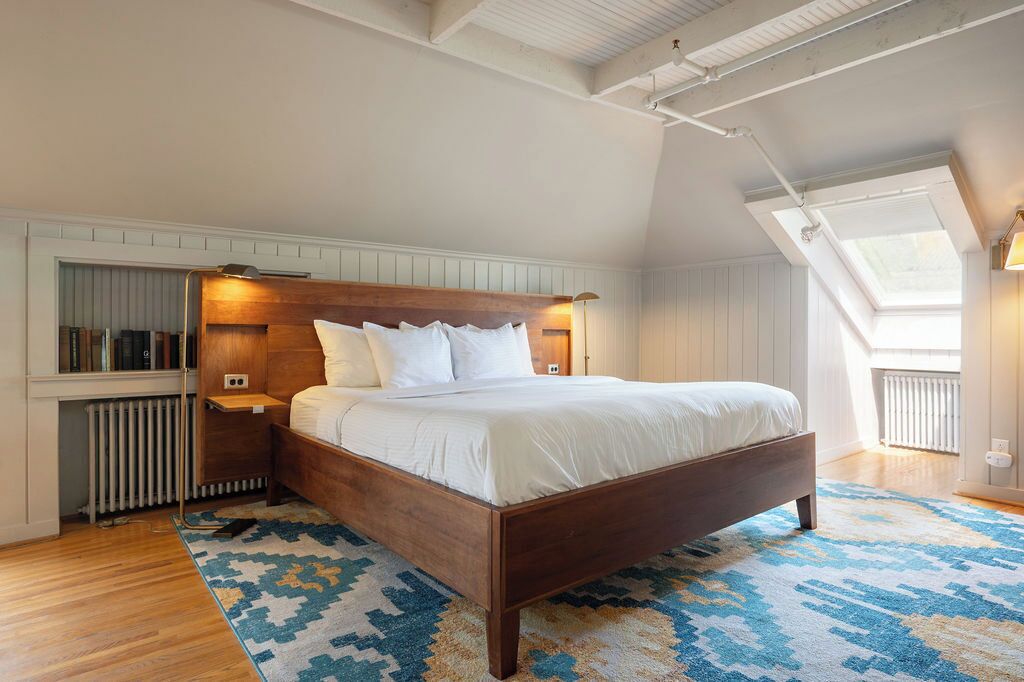 A bedroom with a wooden bed, white bedding, a patterned rug, a skylight, and a radiator.