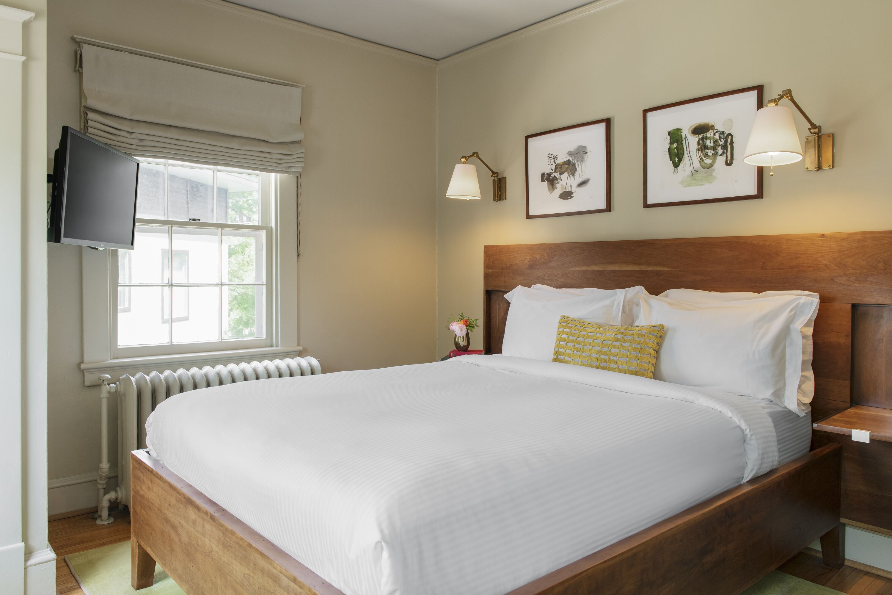 A neatly made bed with white linens, a wooden headboard, and a nightstand with a lamp. A flat-screen TV is mounted on the wall above the bed.