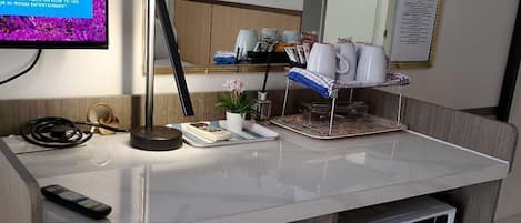Habitación de hotel con escritorio blanco, refrigerador pequeño, televisor, espejo y aire acondicionado instalado en la pared.