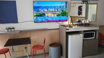 A hotel room with a TV displaying a cityscape, a desk with a chair, a trash bin, and a microwave.