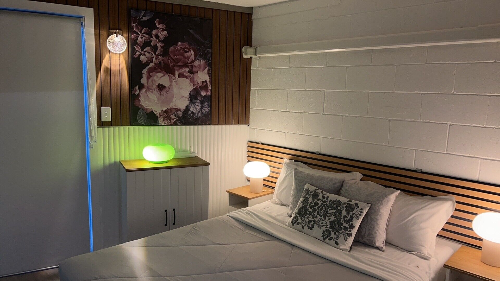 Et soveværelse med en seng, to sengeborde, en kommode, et blomsterbillede og en vægmonteret lampe.