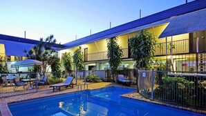 Ein Schwimmbecken mit Liegestühlen und einer überdachten Sitzgruppe, umgeben von einem Gebäude mit flachem Dach und großen Fenstern.