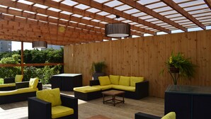 Un patio al aire libre con suelo de madera, mobiliario amarillo y negro, y estructura de techo de madera.