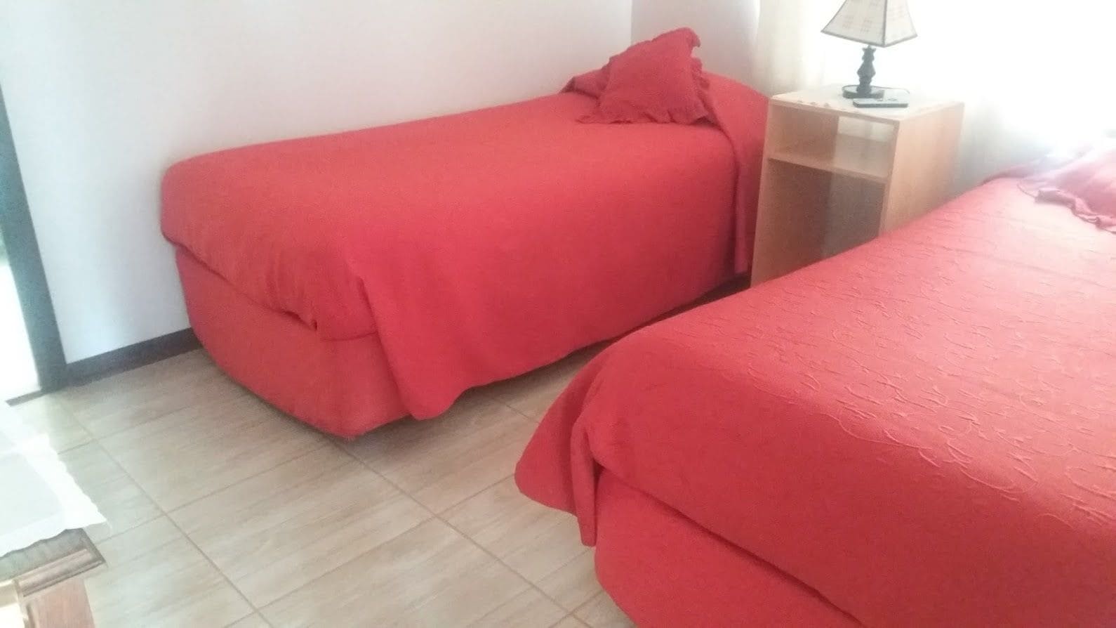 Two single beds with red bedding in a room with a tiled floor and a bedside table with a lamp.