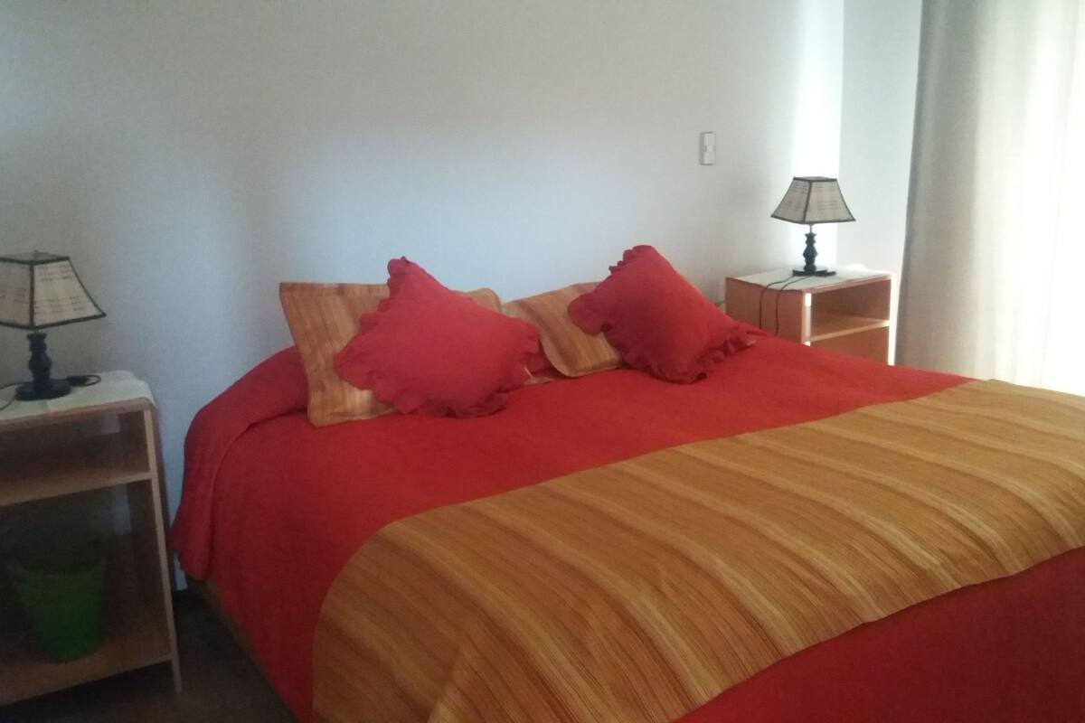 A bed with a red and beige striped bedspread, two red pillows, and a wooden nightstand with a lamp.