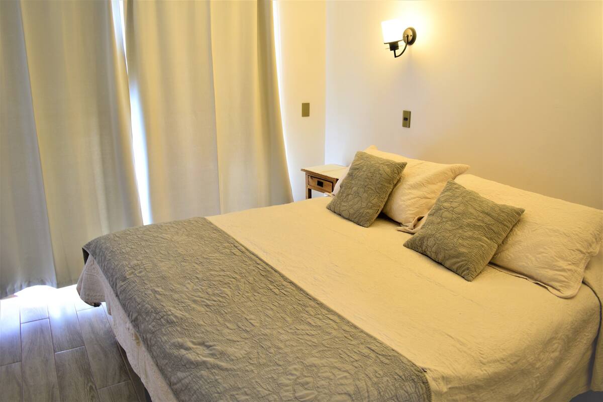 A neatly made bed with a patterned bedspread and two pillows, a bedside table, and a wall-mounted light fixture.