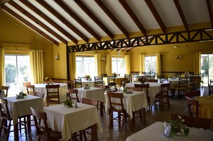 Un comedor con varias mesas y sillas, vigas de madera en el techo y ventanas amplias con cortinas.