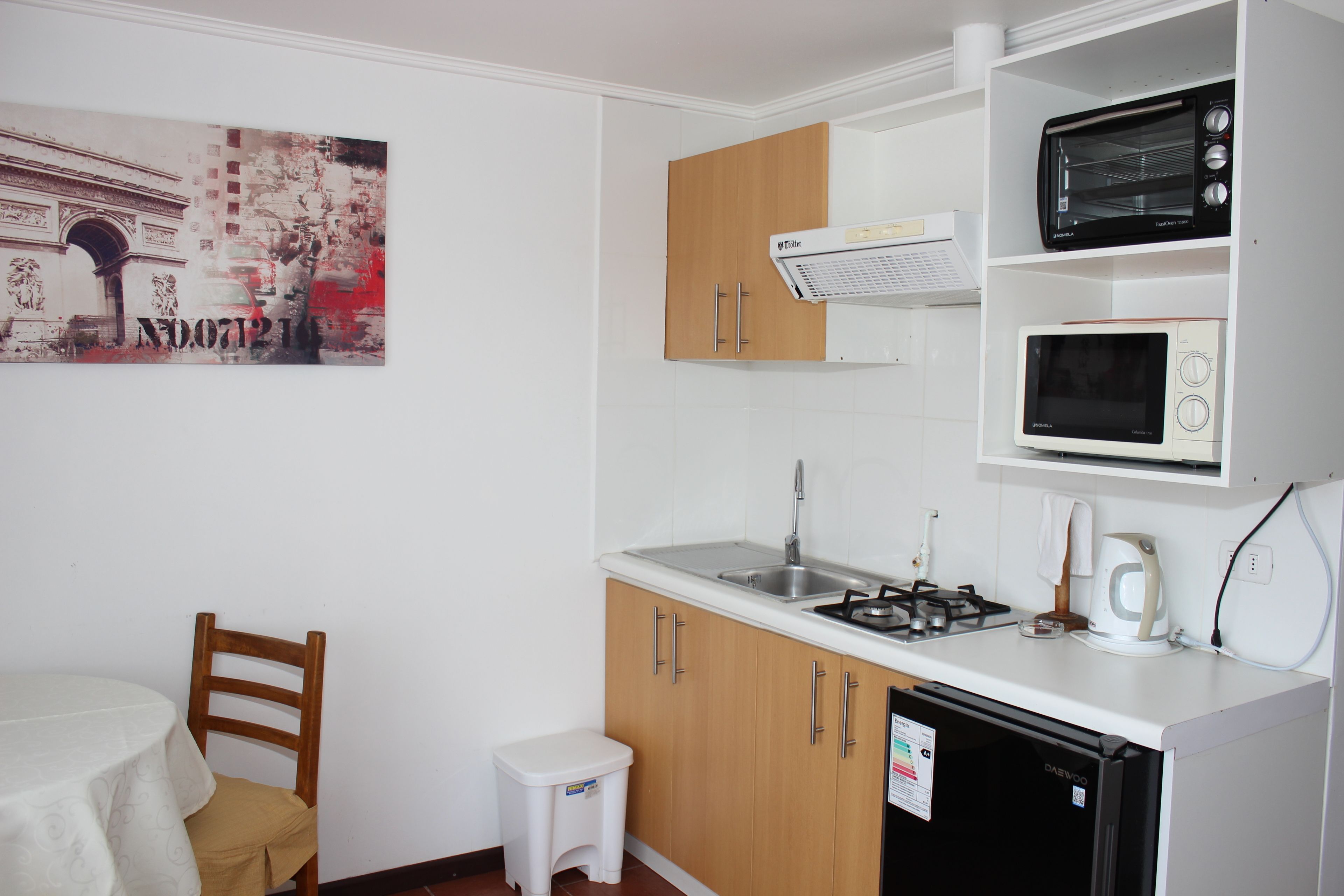 A compact kitchen with a sink, stove, and microwave. There is a small white stool and a wooden chair.